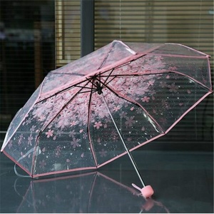 Transparenter Regenschirm mit Blumenmotiv für Kinder mit rosa Rand auf einem Tisch vor einem Fenster mit geschlossenen Vorhängen