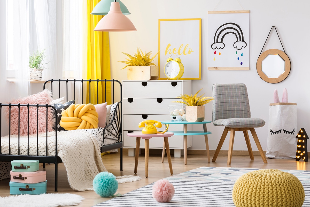 Gelber Pouf, grauer Stuhl und Gitterbett in einem farbenfrohen Kinderzimmer mit Poster und Wanddekorationen