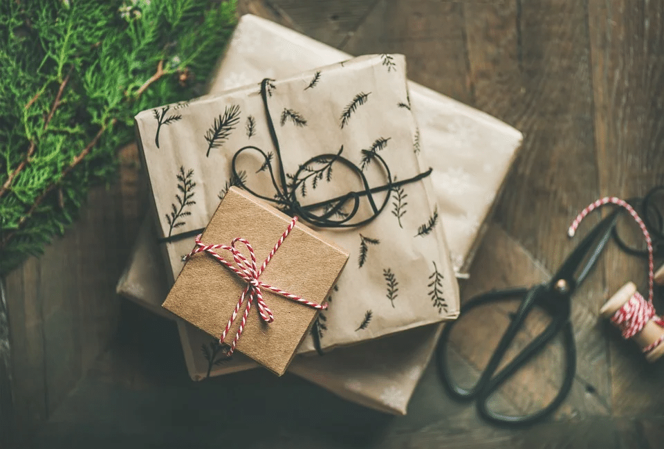 3 graue und braune Geschenkpakete, mit Geschenkband umwickelt, auf einem braunen Holzboden mit einem Stemmeisen und einem Tannenzweig im Hintergrund