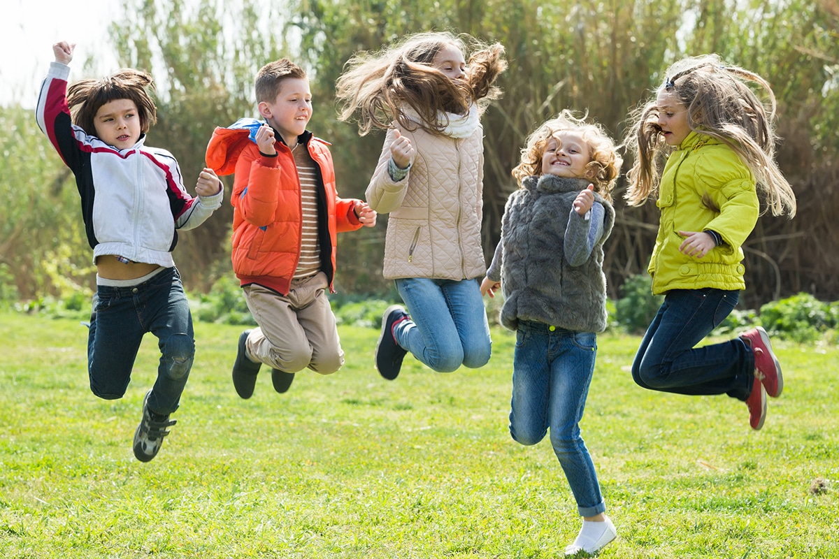 Eine Gruppe fröhlicher Kinder springt im Gras im Freien. Es sind 2 Jungen und 3 Mädchen. Im Hintergrund stehen Bäume
