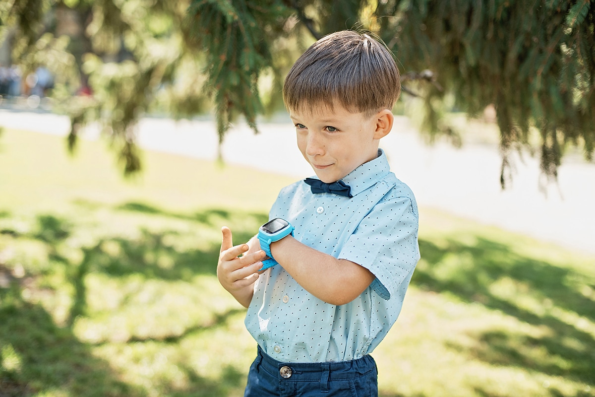 Ein Junge spricht in seine Armbanduhr. Er trägt eine blaue Armbanduhr, ein blaues Hemd und ist im Freien in der Sonne, mit Bäumen im Hintergrund