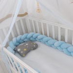 Blau geflochtener Bettumrandung in einem weißen Babybett mit weißer Bettwäsche und einem transparenten Vorhang