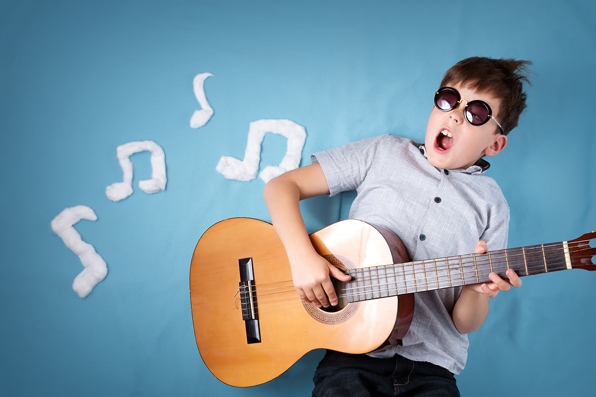 Ein kleiner Junge auf blauem Hintergrund, der Gitarre spielt. Der Junge trägt eine schwarze Brille, ein graues kurzärmeliges Hemd und eine braune Holzgitarre. Im Hintergrund sind weiße Noten zu sehen