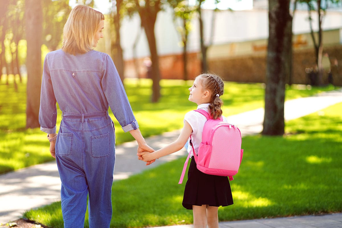 Eine Mutter geht mit ihrer Tochter in einen Park. Es ist sonnig, die Mutter trägt blaue Jeans, die Tochter einen rosa Kinderrucksack