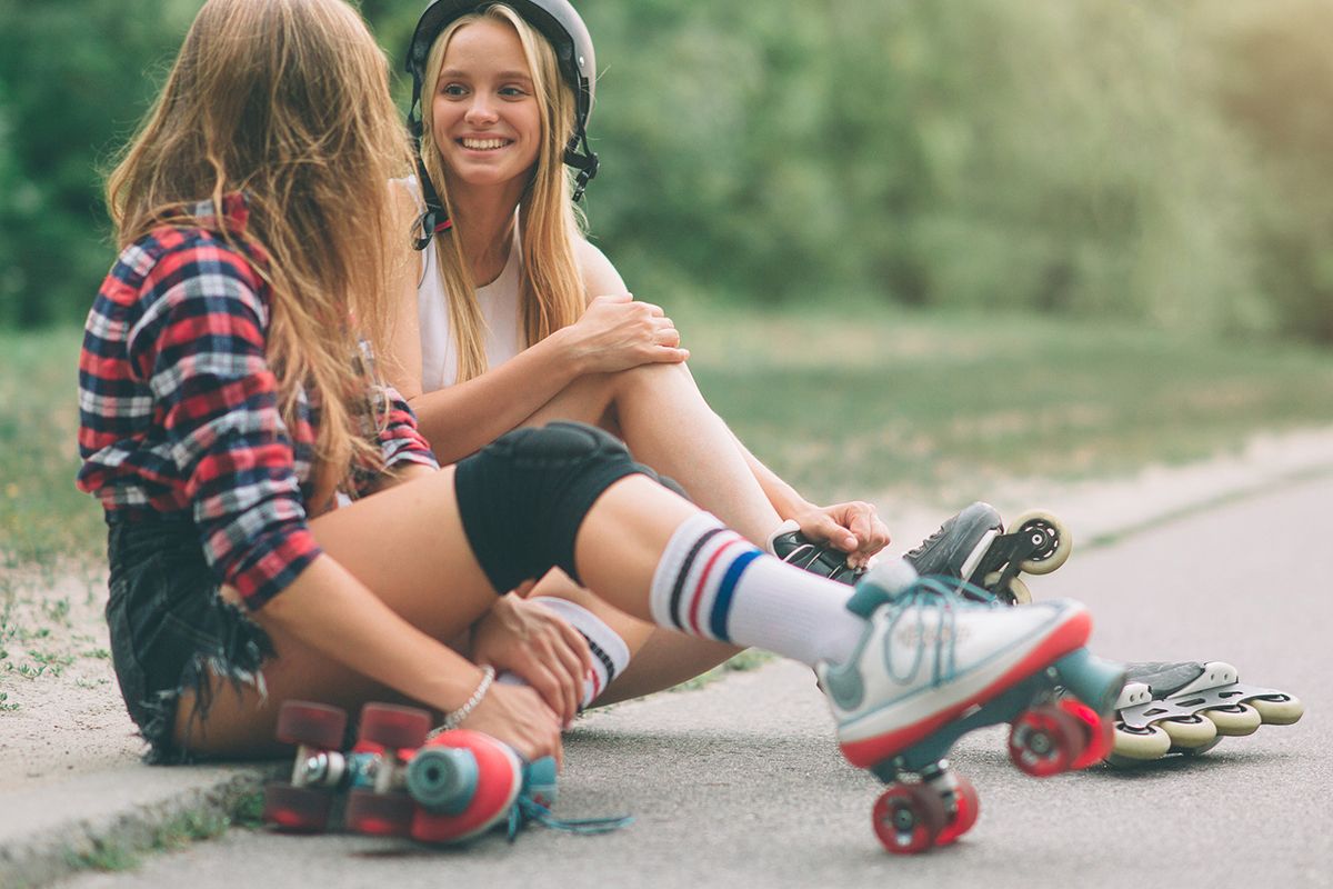 Zwei junge Mädchen sitzen am Straßenrand. Ein Mädchen trägt Rollschuhe, das andere Inlineskates und einen Helm.