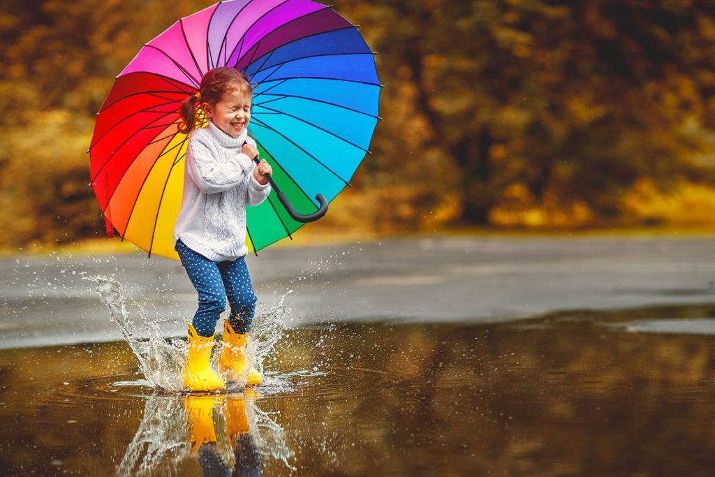 Une petite fille qui tient un parapluie pour enfant de couleur arc-en-ciel. Elle porte un pull beige et des bottes de pluie jaunes. Elle se tient dans une grande flaque d'eau