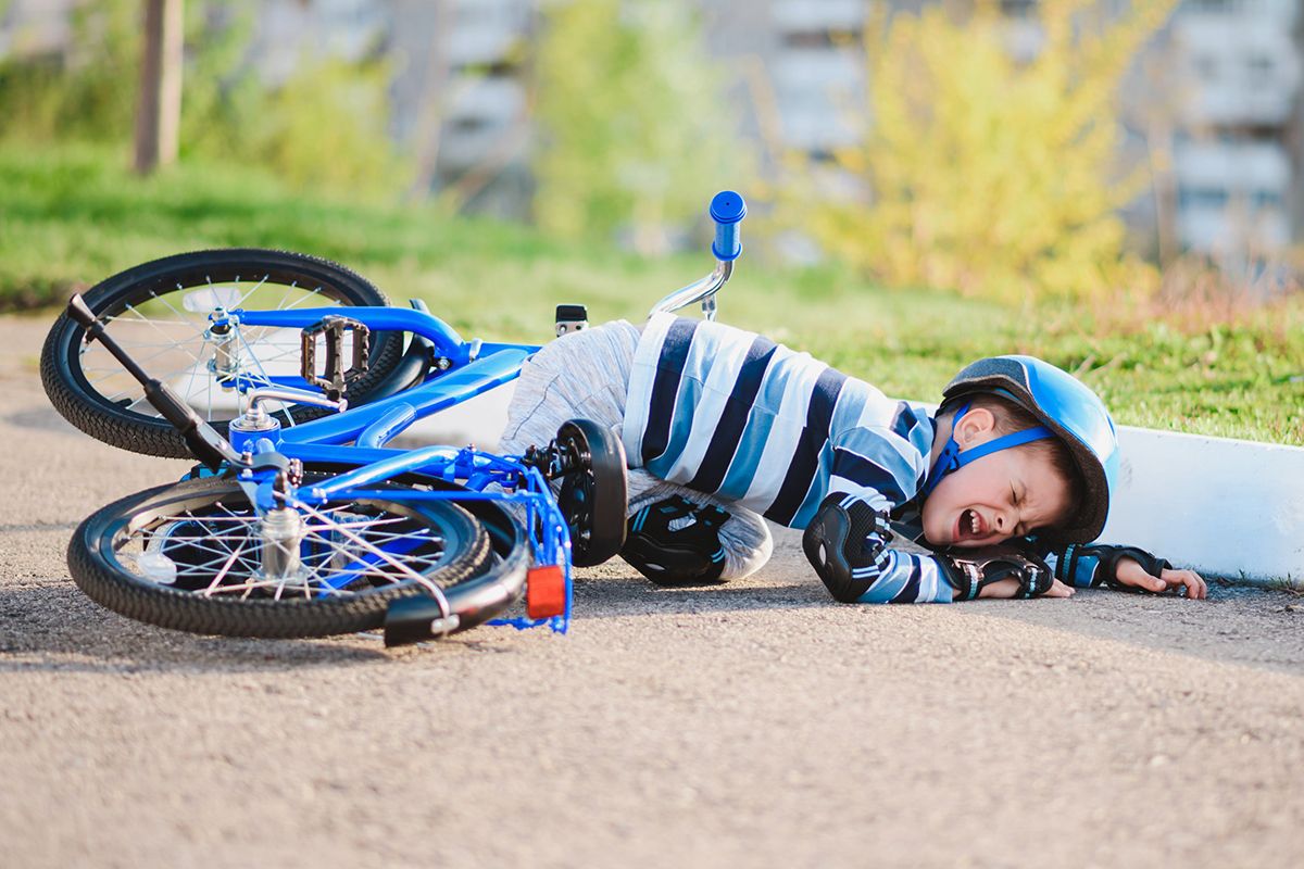 Ein kleines Kind, das von seinem blauen Fahrrad gefallen ist. Es weint und trägt einen schwarzen Fahrradhelm, Knieschützer und Schutzhandschuhe