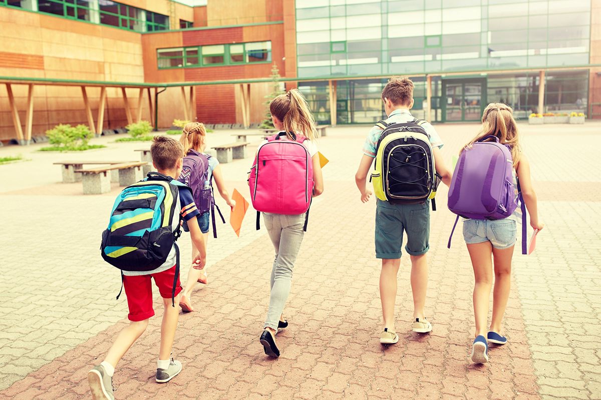 Eine Gruppe von fünf Kindern, die auf dem Weg zur Schule sind. Jedes Kind trägt eine farbige Schultasche oder einen farbigen Kinderrucksack