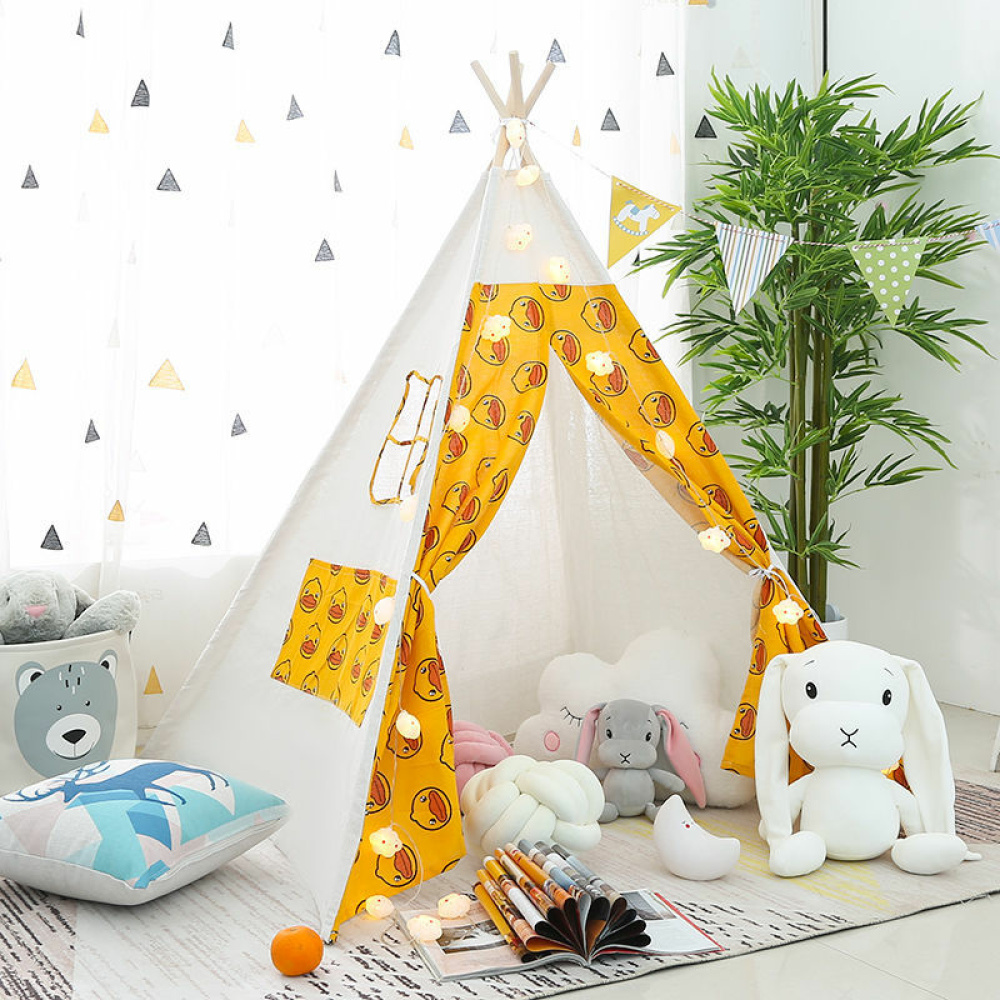 Ein Tipi im indianischen Stil für Kinder in einem Kinderzimmer. Es ist weiß mit gelben Türflügeln mit niedlichen Motiven. Es steht auf einem Teppich auf dem Boden. Neben ihm stehen bunte Cousins, Kuscheltiere und ein Strauch dahinter.