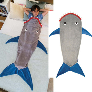 Das Foto ist in zwei Teile geteilt. Der erste Teil zeigt ein Kind, das in einem Bett liegt und in einen Schlafsack in Form eines grauen Hais mit blauen Flossen und einem roten Mund gesteckt wird. Der zweite Teil des Bildes zeigt diesen Schlafsack vor einem weißen Hintergrund.